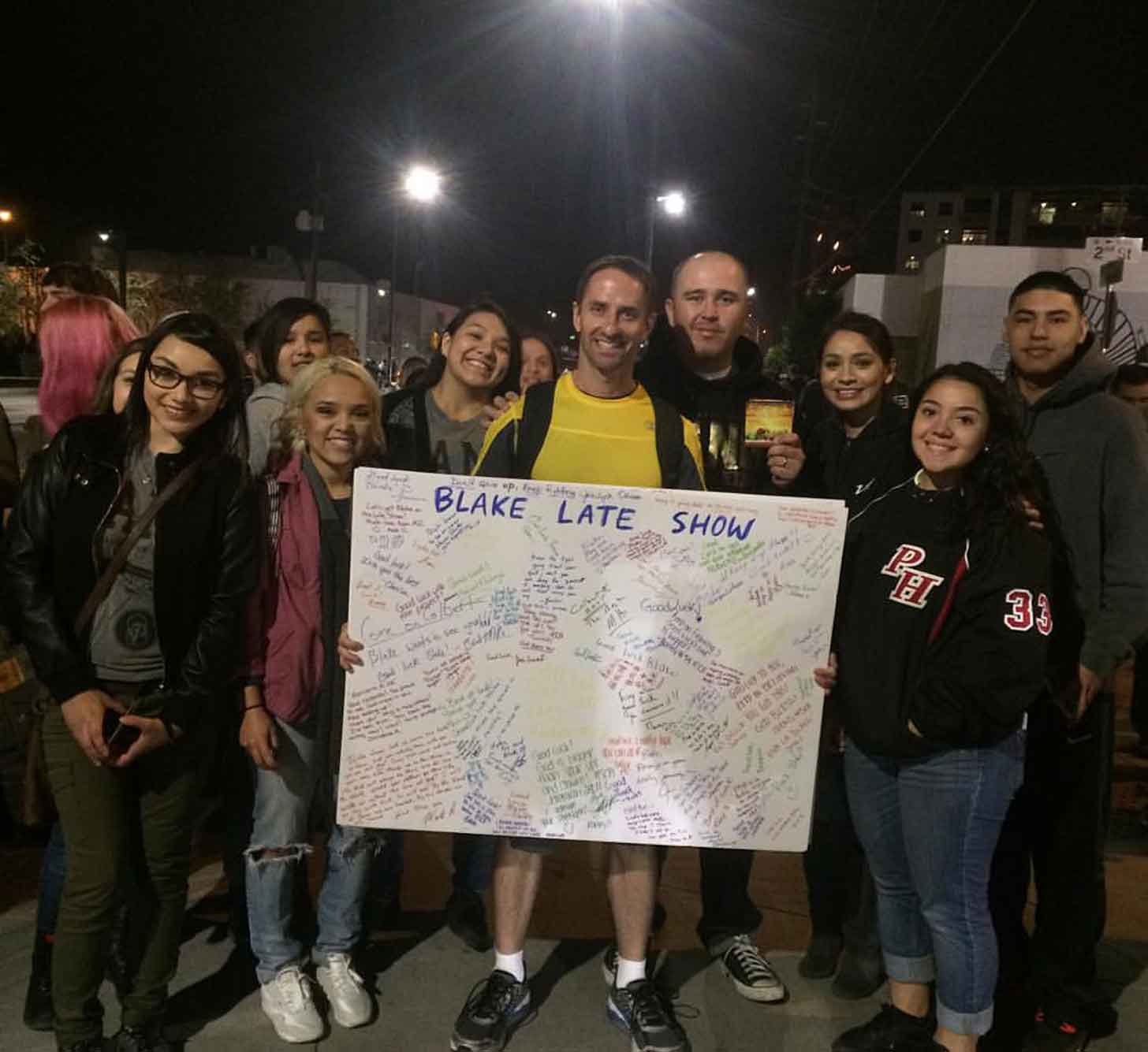 A man named Ron Blake holds up a poster board with colorful signatures. He is surrounded by smiling strangers.