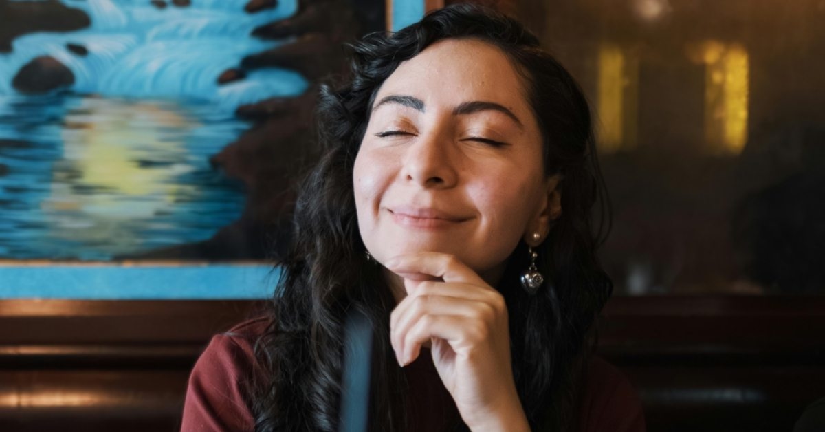 A contemplative young woman smiles in a restaurant, resting her finger on her chin.