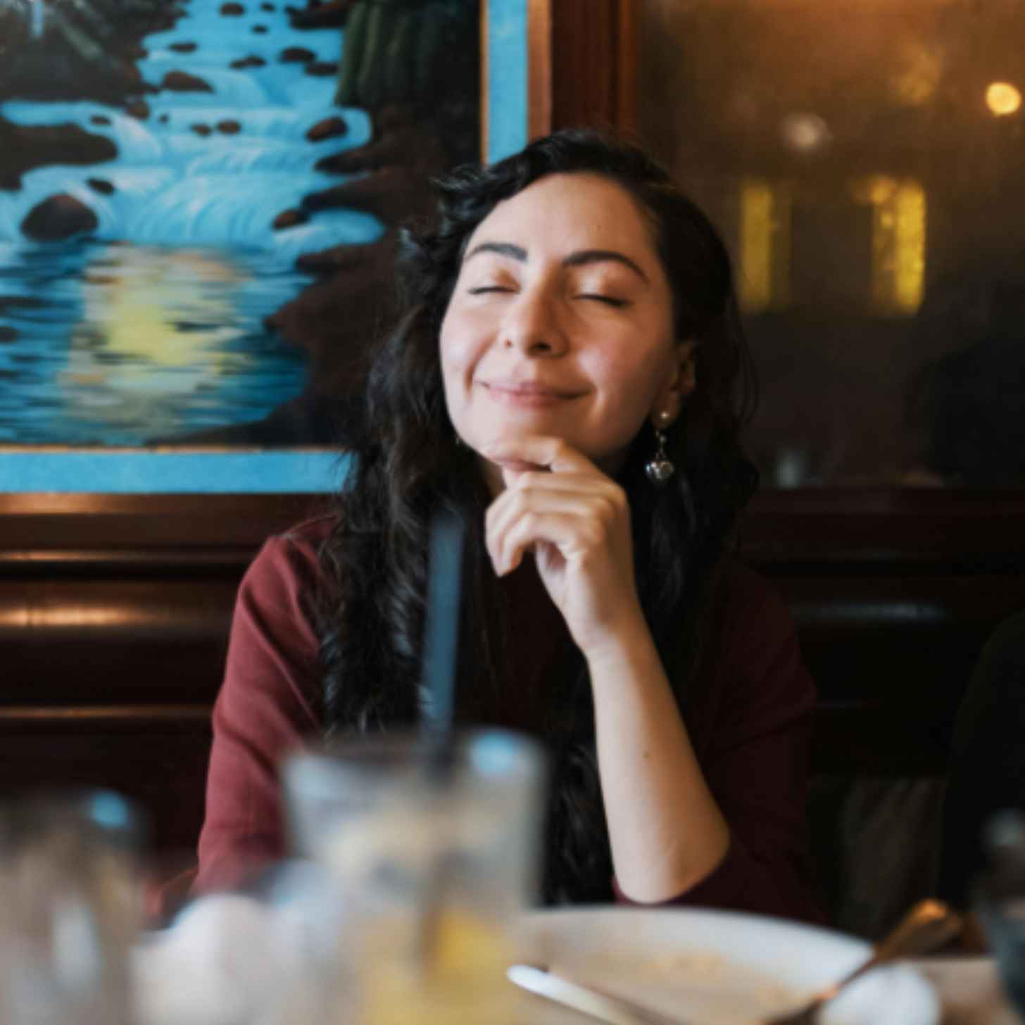 A contemplative young woman smiles in a restaurant, resting her finger on her chin.