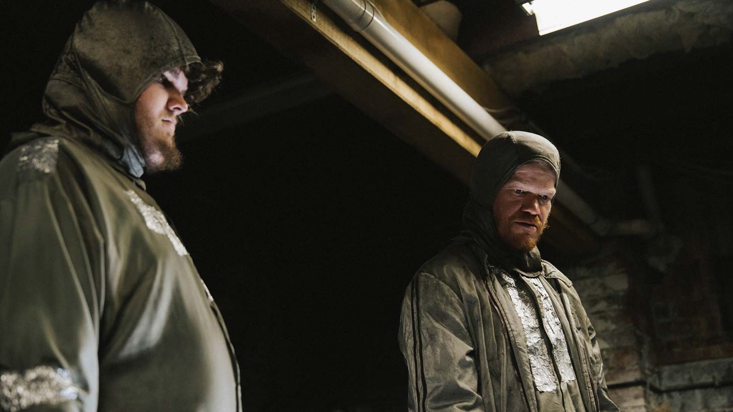 Aidan Delbis and Jesse Plemons wear PPE in a dark basement in "Bugonia"