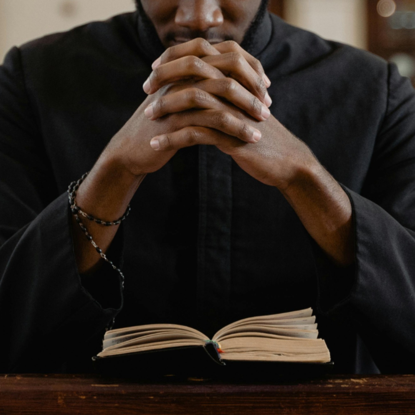 A young Black man kneels at a church pew with a rosary and bible in front of him and his hands folded in prayer