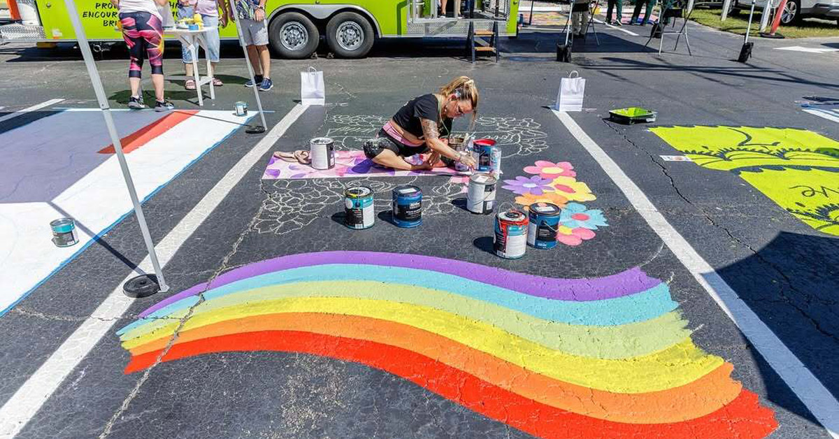 A woman with blond hair paints a rainbow on a concrete parking space