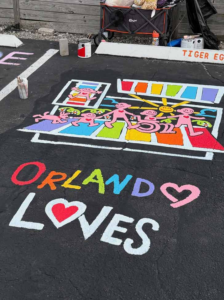 A painted mural shows pink people walking across a rainbow crosswalk that reads "Orlando Loves" 