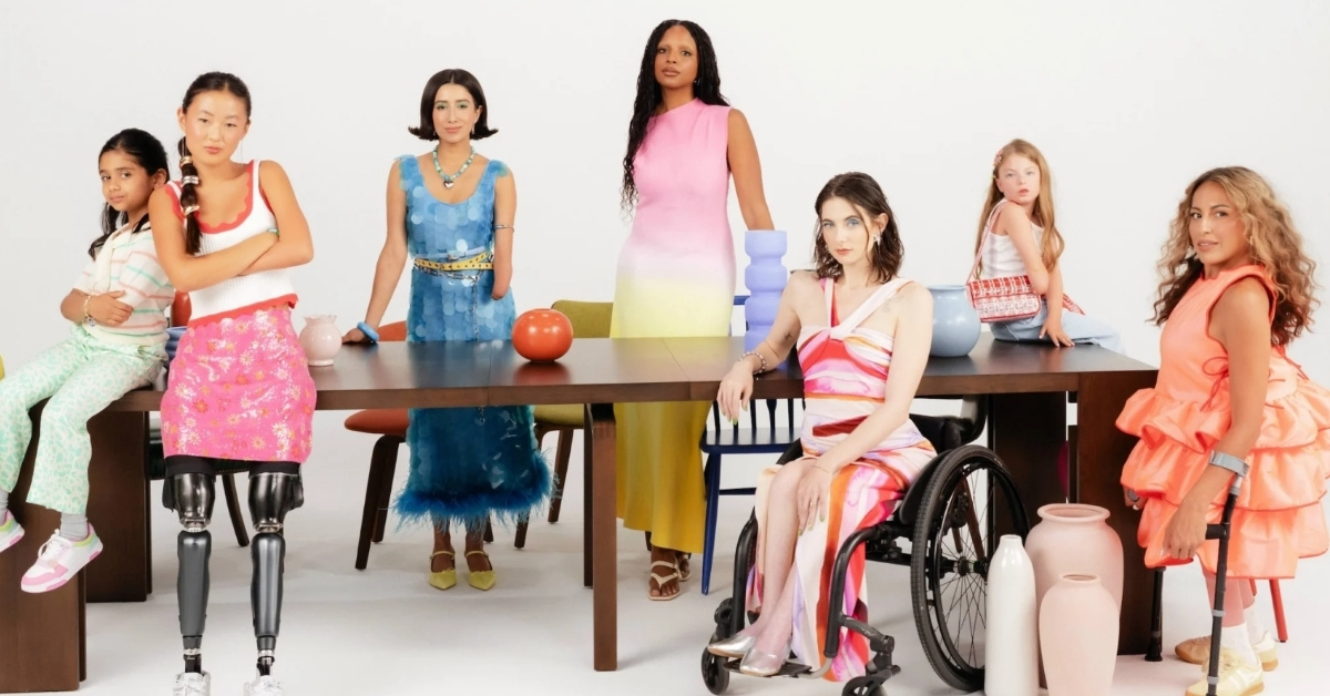 A group of women of assorted ages, races, and disabilities stand around a table for a fashion photoshoot. They are wearing bright, fashionable outfits.