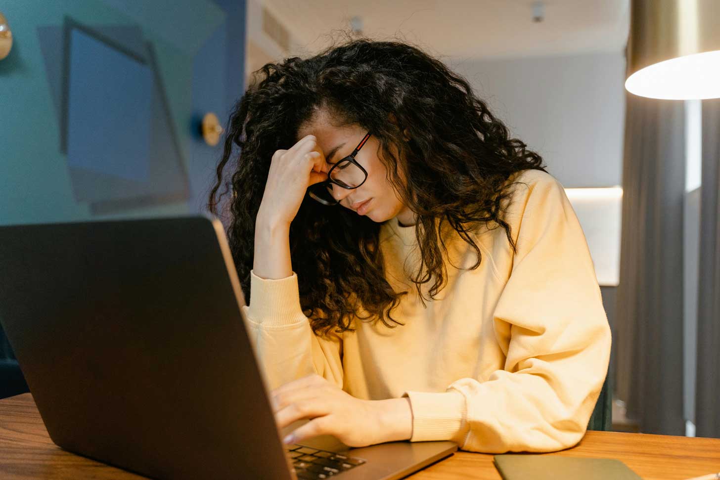 A woman with long, dark curly hair, hunches over a laptop, with her hand pinching the bridge of her nose