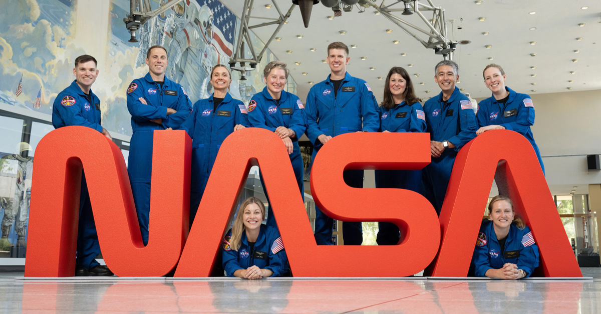 A group of ten astronauts in blue space suits — 4 men and 6 women — pose with red NASA letters 