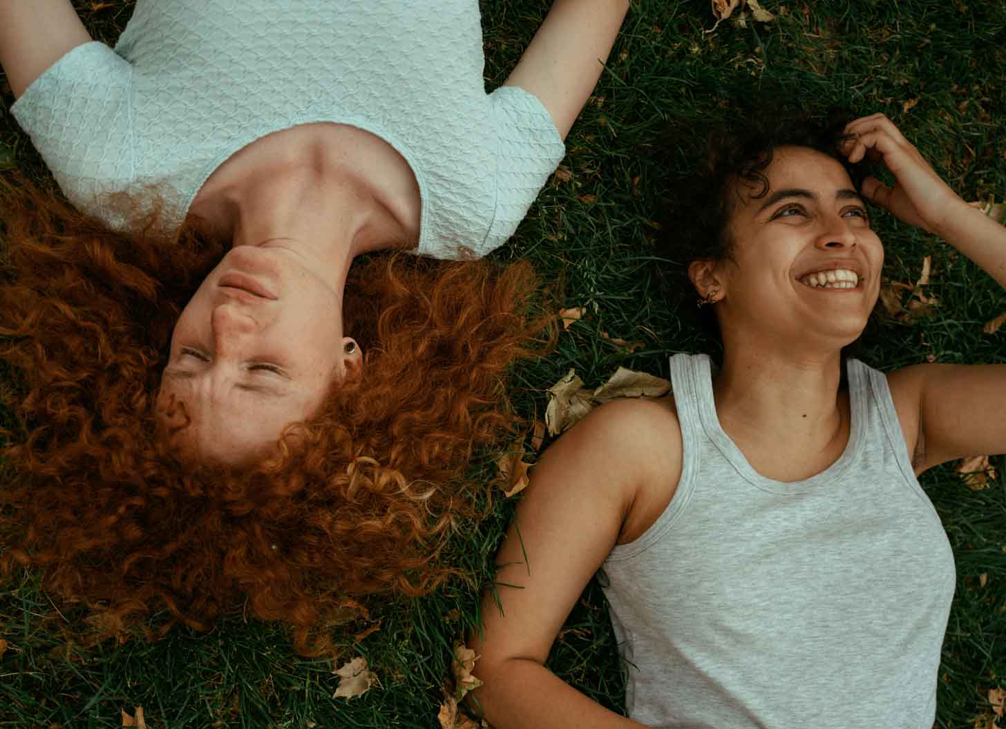 A young woman with long, red curly hair lays next to a young woman with dark curly hair in the grass, smiling