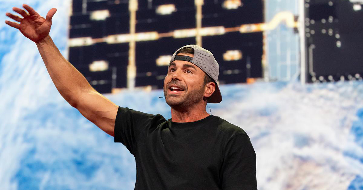 Mark Rober speaks on stage at TED in front of a photo of a satellite in space. He wears a black t-shirt and backwards baseball cap