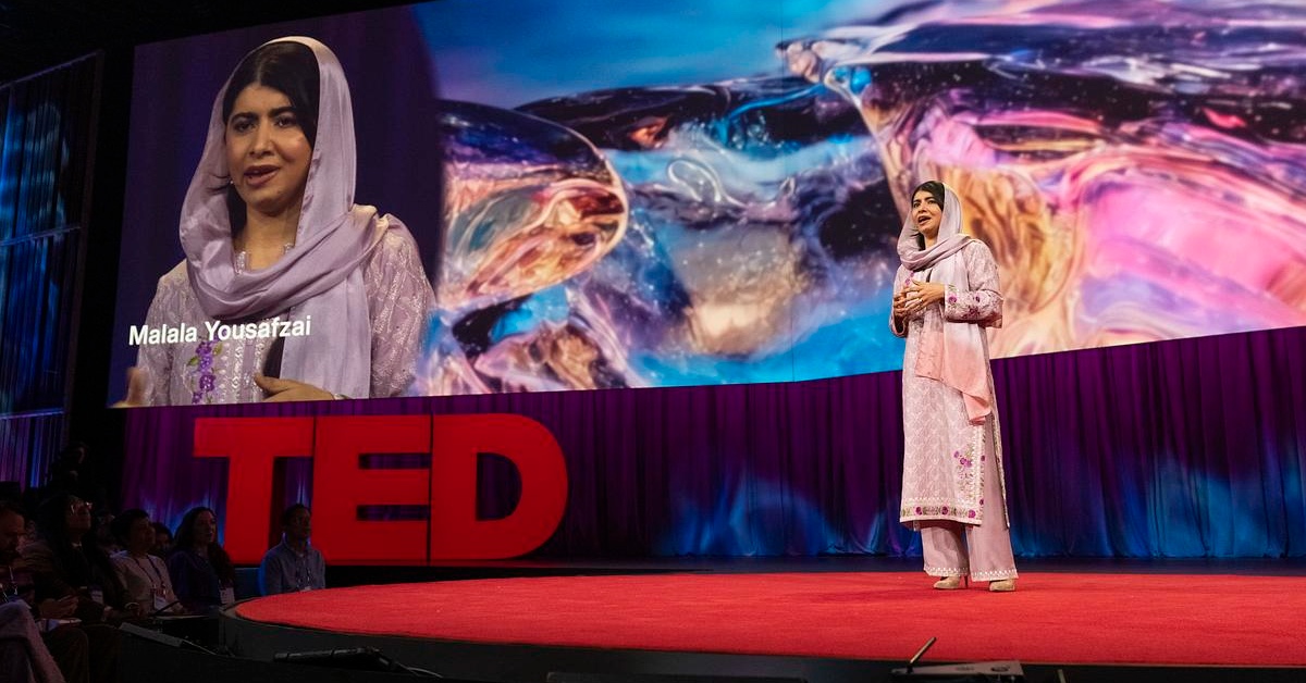 Malala, dressed in pink, stands on stage for TED Talk, standing on red carpet with the TED logo in the background.