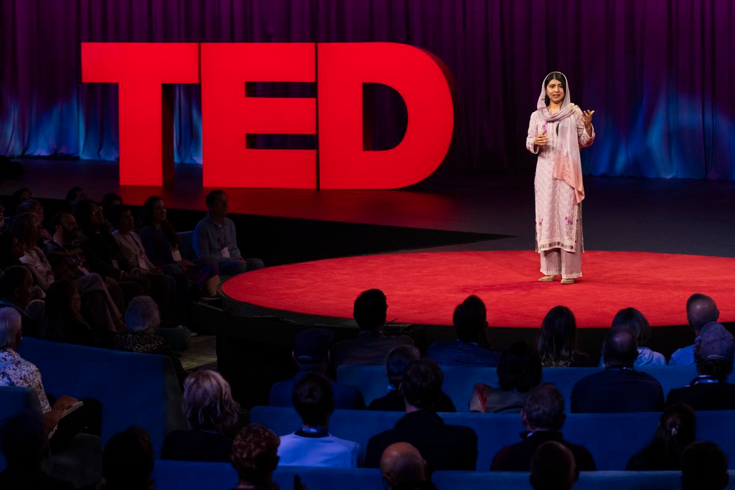 Malala, dressed in pink, stands on stage for TED Talk, standing on red carpet with the TED logo in the background.
