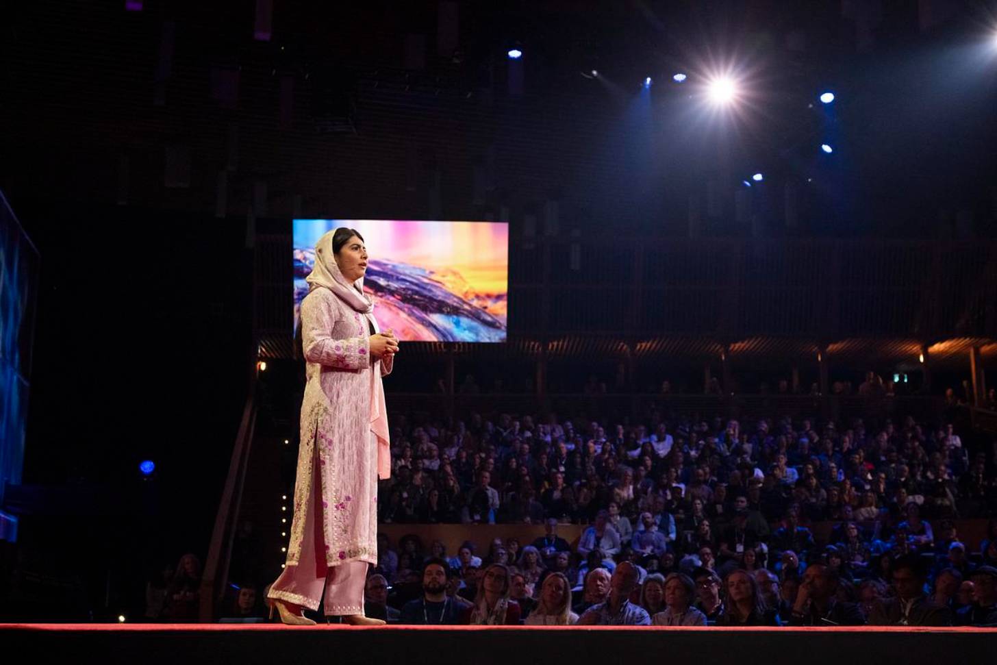 Malala, dressed in pink, stands on stage for TED Talk