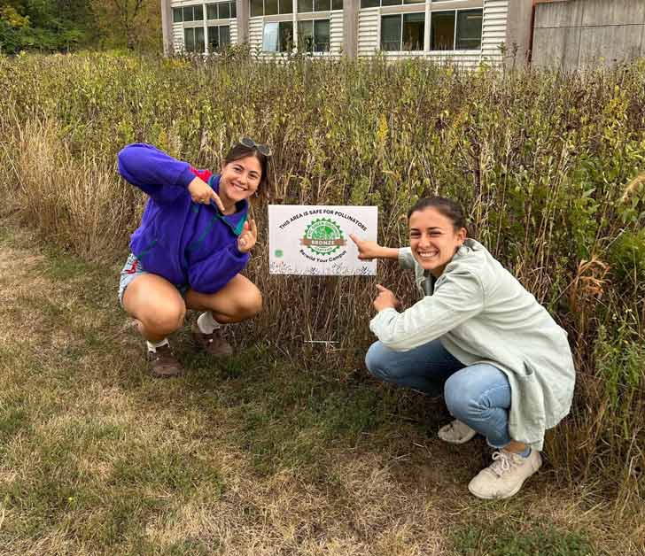 two female college students crouching next to a garden, smiling