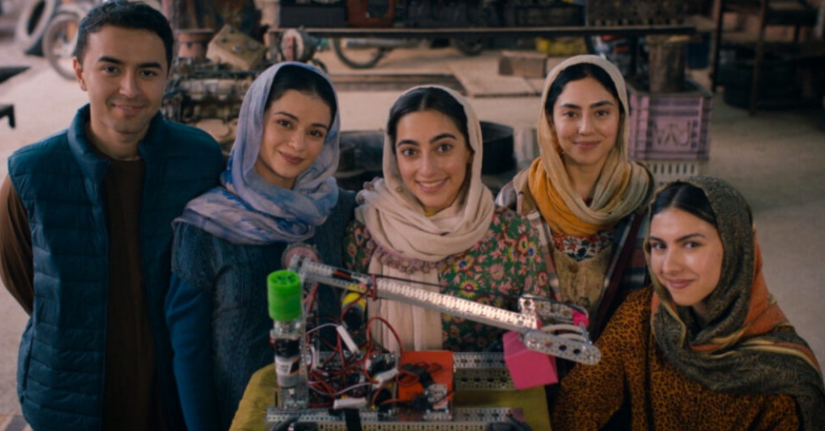 A team of young women, and one man, smile around a small robot.