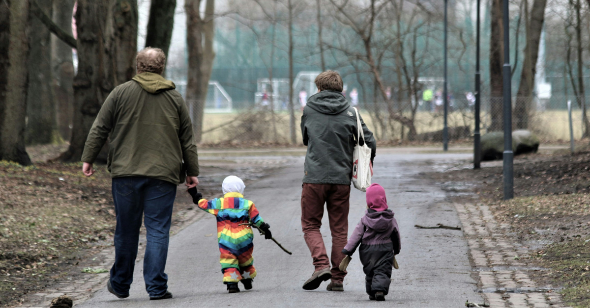 Two adults walk along a path with two toddlers. Only their backs are visible