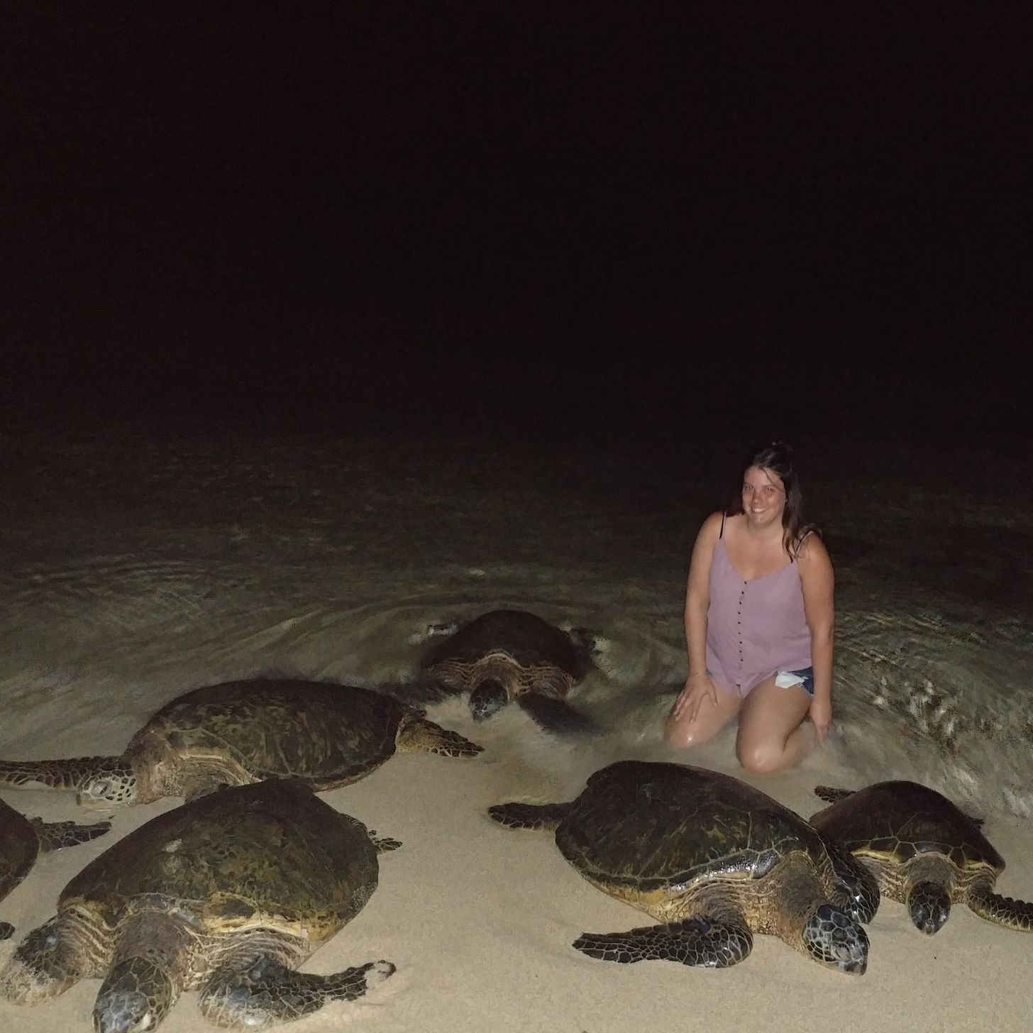 A woman smiles on a beach at night, surrounded by green sea turtles