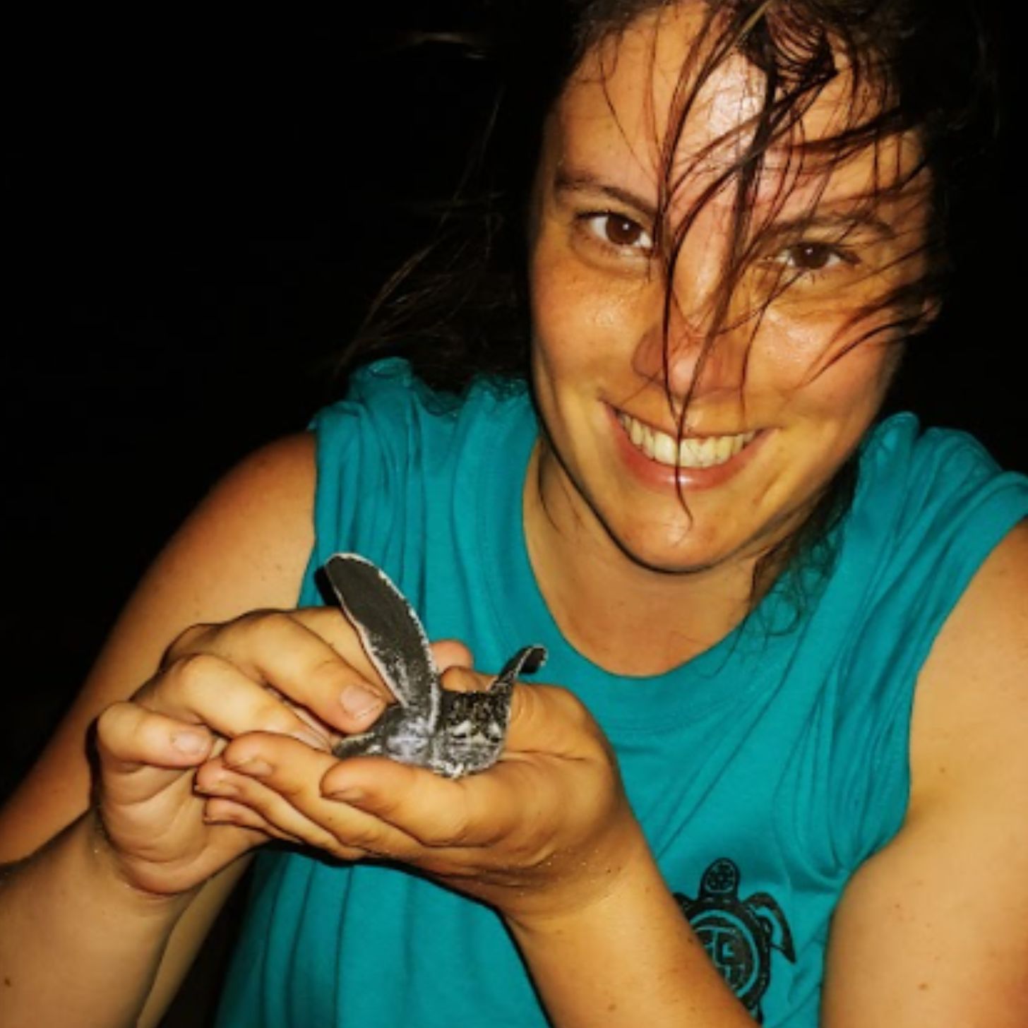 In a closeup, a smiling woman holds up a baby leatherback turtle on a beach at night.