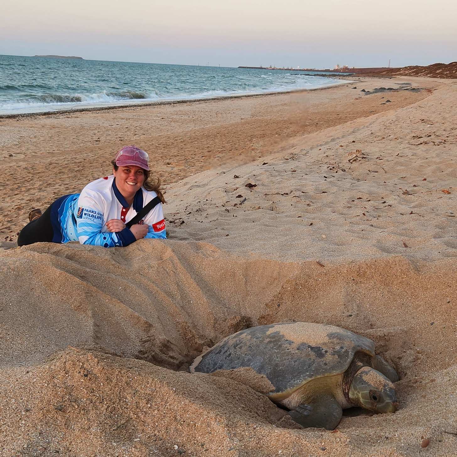 A woman smiles as she lies in the sand beside a large flatback sea turtle on a beach.