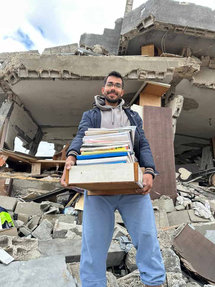 A man holds a stack of books in the rubble of Gaza