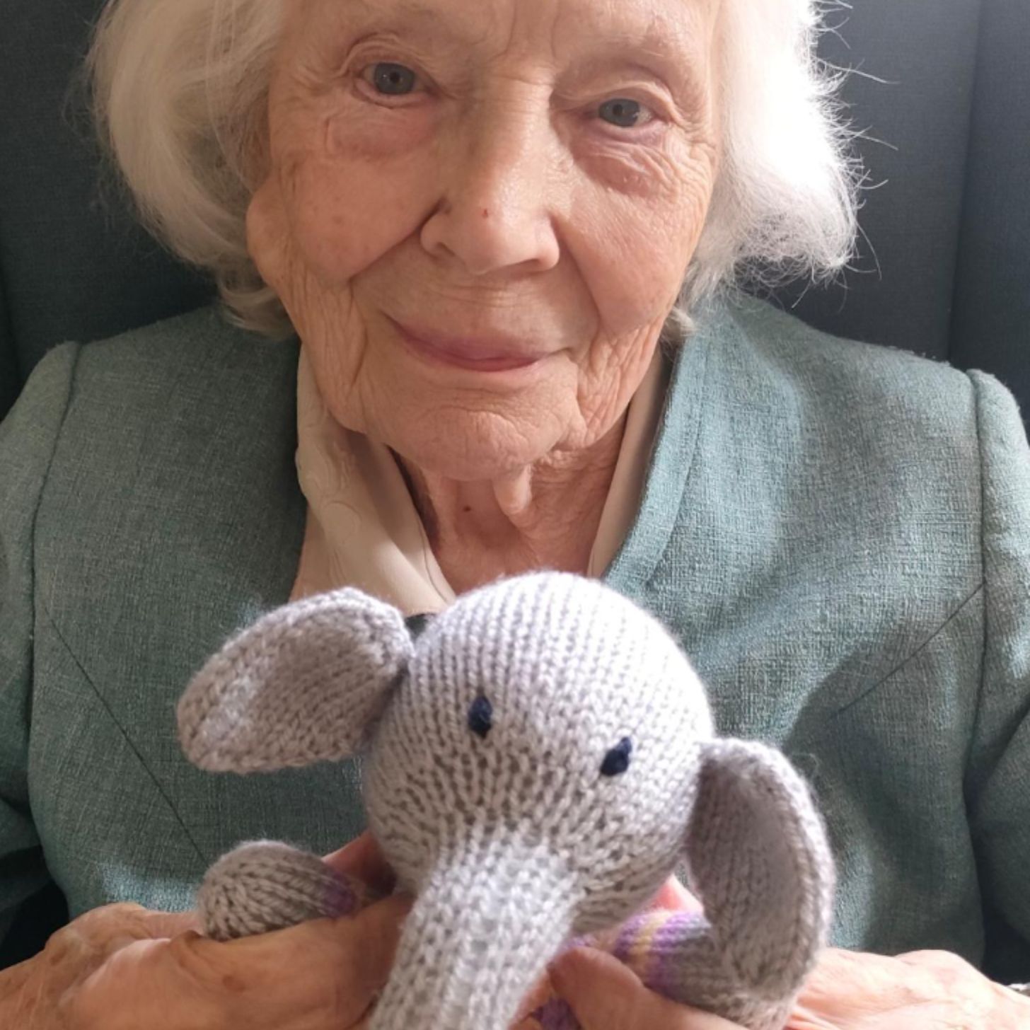 An elderly woman smiles softly as she holds up a handmade elephant plushie
