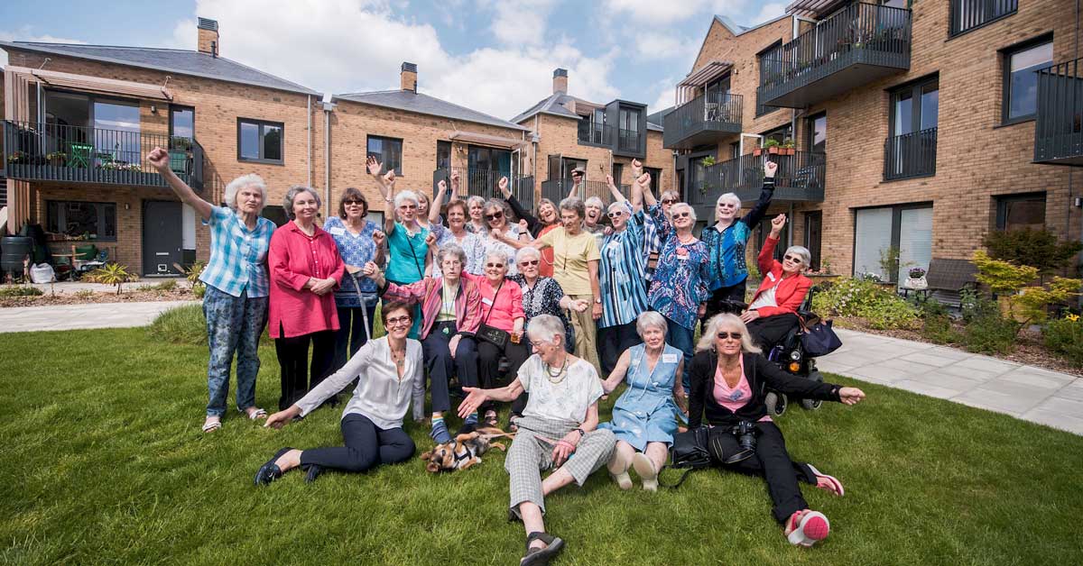 A group of 26 elderly women smiling with their arms out in front of a brick apartment complex in North London