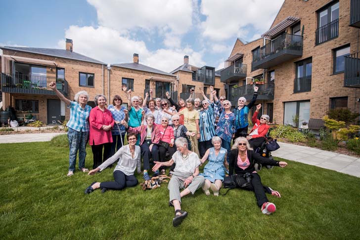 A group of 26 elderly women smiling with their arms out in front of a brick apartment complex in North London