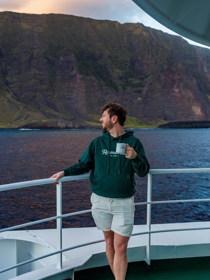 A man, Jake Rosmarin, stands on the helm of a cruise ship, coffee mug in hand, looking out at the ocean