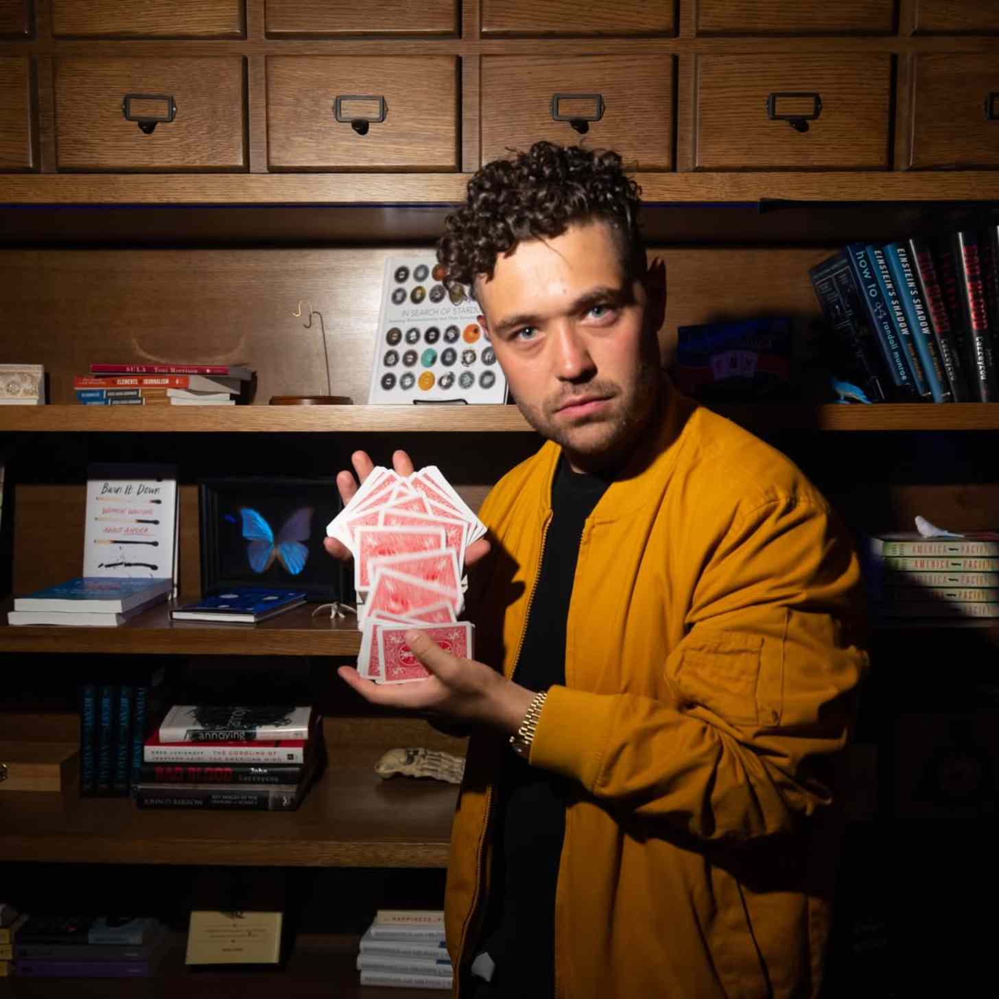 Magician John Stessel holds up a deck of cards in front of a bookshelf
