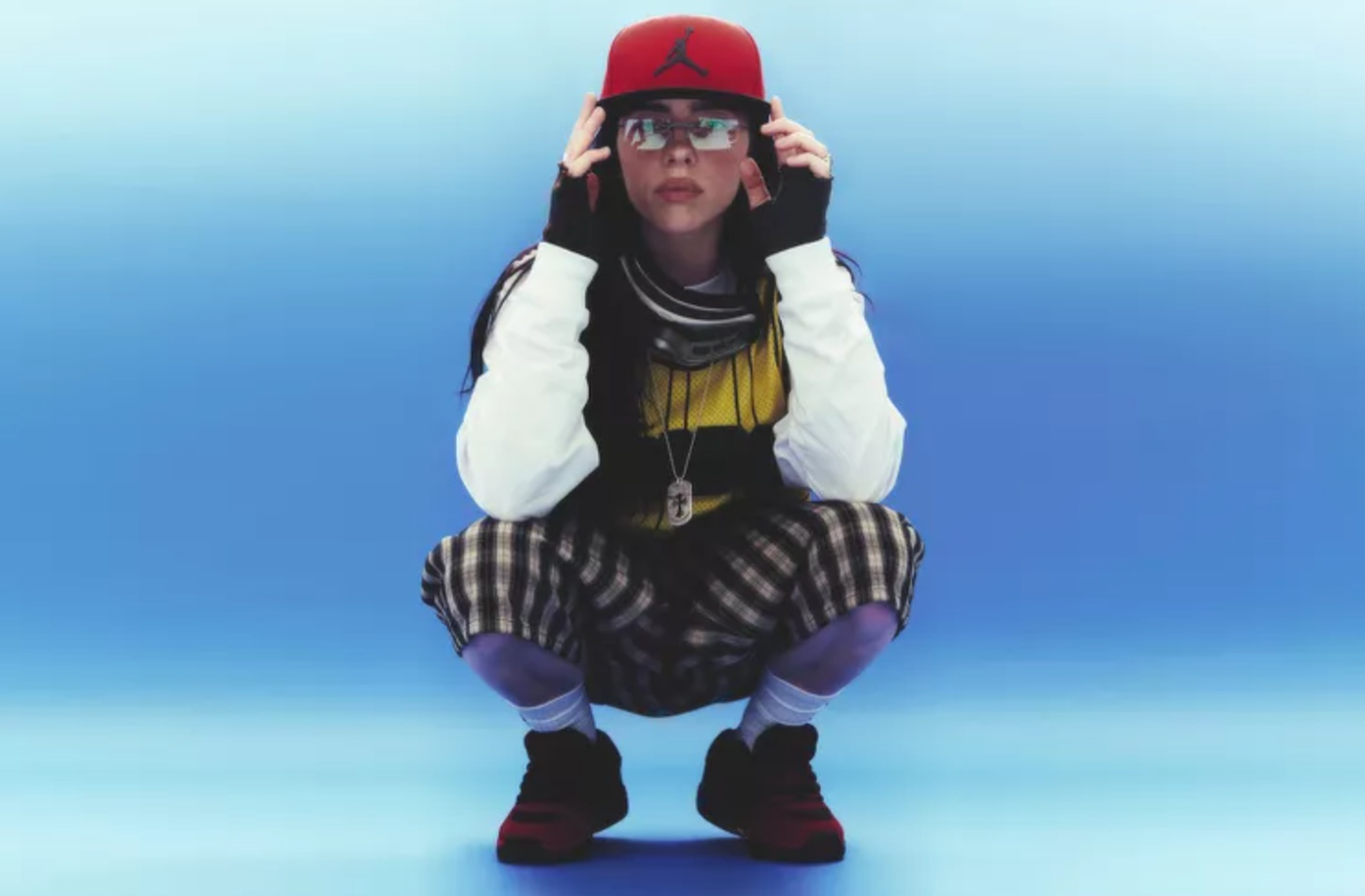 Billie Eilish squats down in a post for a promotional image for her Hit Me Hard and Soft Tour. There is a faded blue backdrop behind her