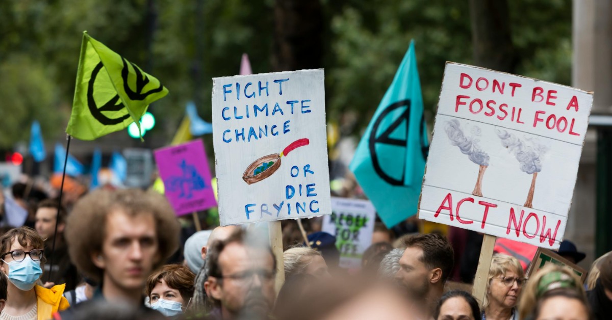 A crowd protests climate change issues with colorful signs. One says: Fight Climate Change or Die Frying. Another says: Don't Be a Fossil Fool, Act Now!