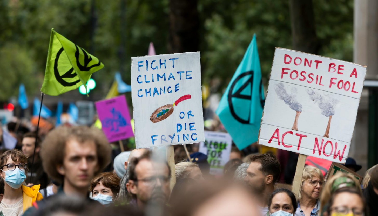A crowd protests climate change issues with colorful signs. One says: Fight Climate Change or Die Frying. Another says: Don't Be a Fossil Fool, Act Now!