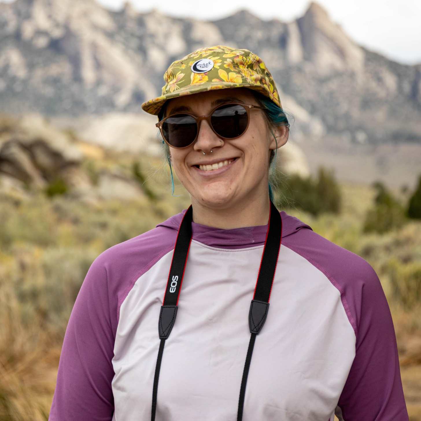 A young woman with blue hair and a floral hat wears sunglasses in front of a mountain range