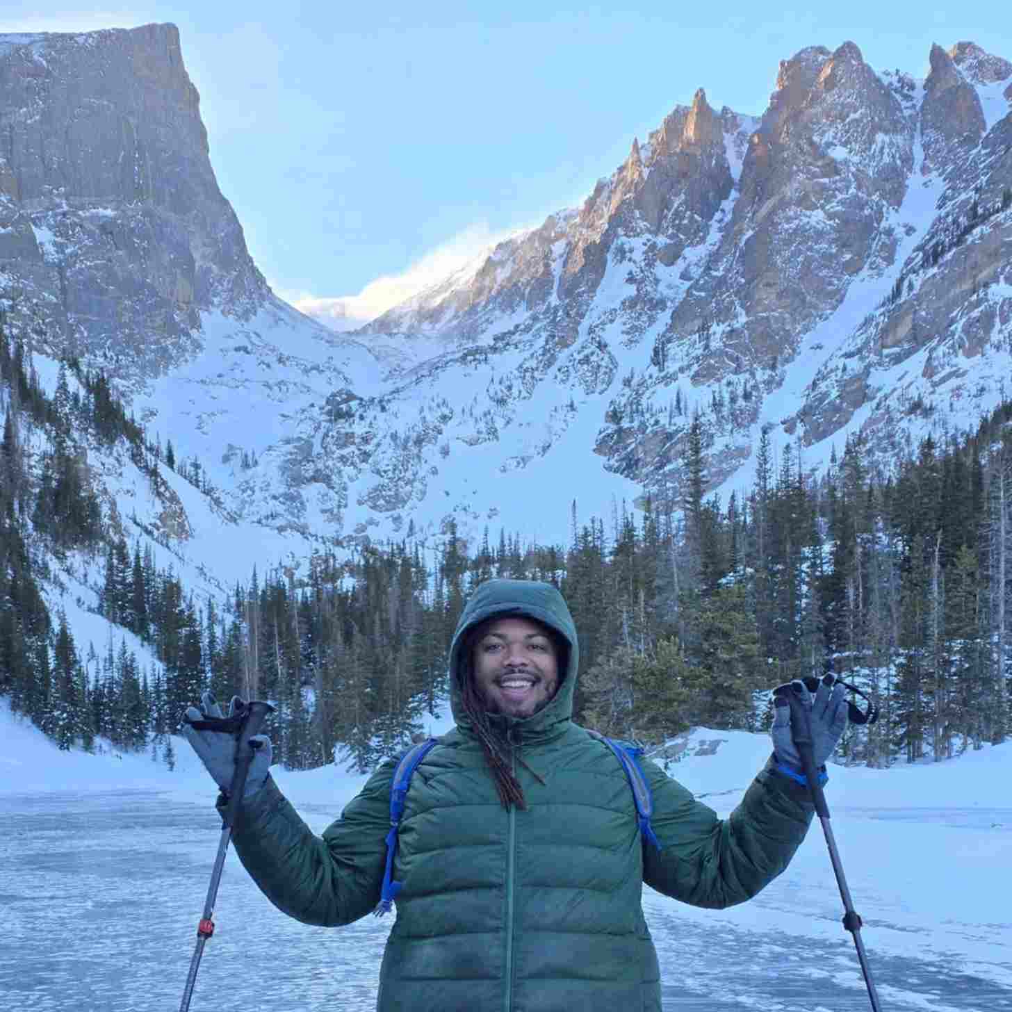 A Black man wears a green puffer coat in front of a snowy mountain, smiling