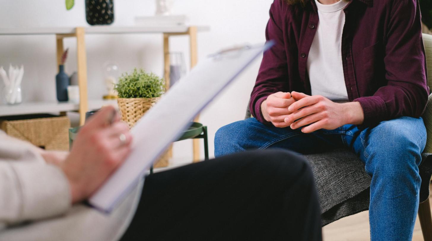 A man sits with his hands folded in his lap in a therapy office