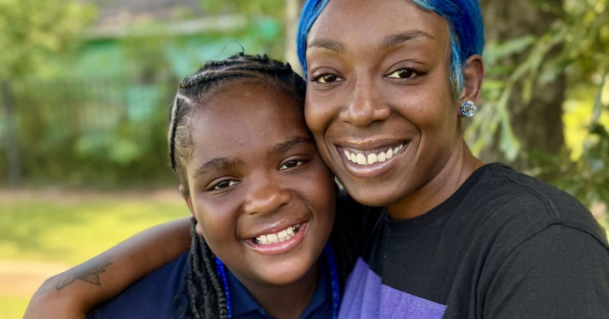 A Black woman with blue hair smiles as she embraces a younger woman