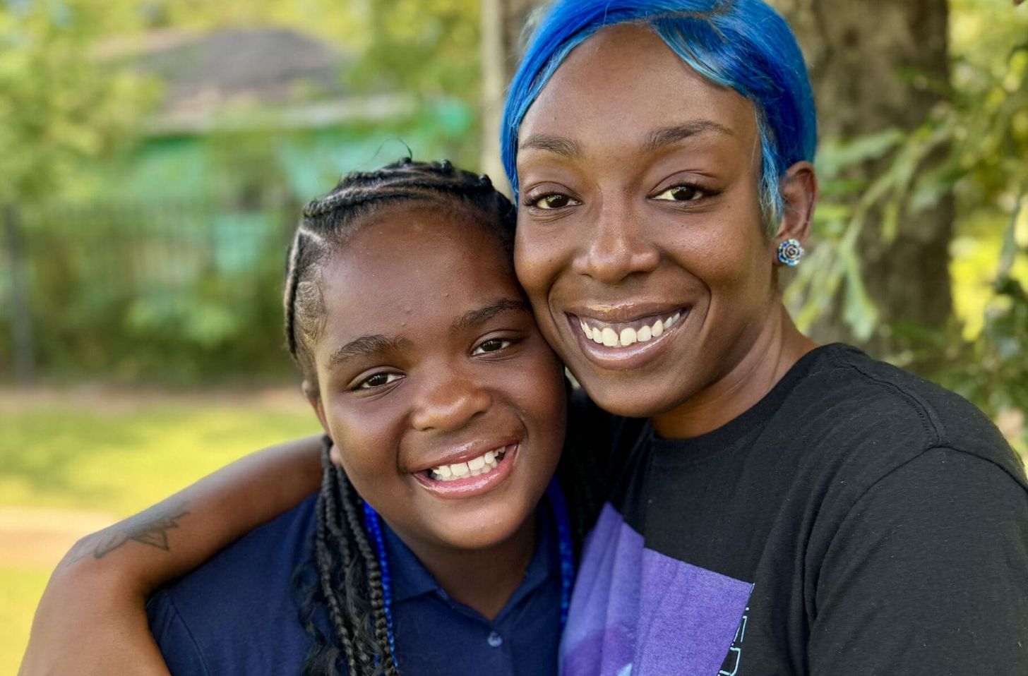 A Black woman with blue hair smiles as she embraces a younger woman