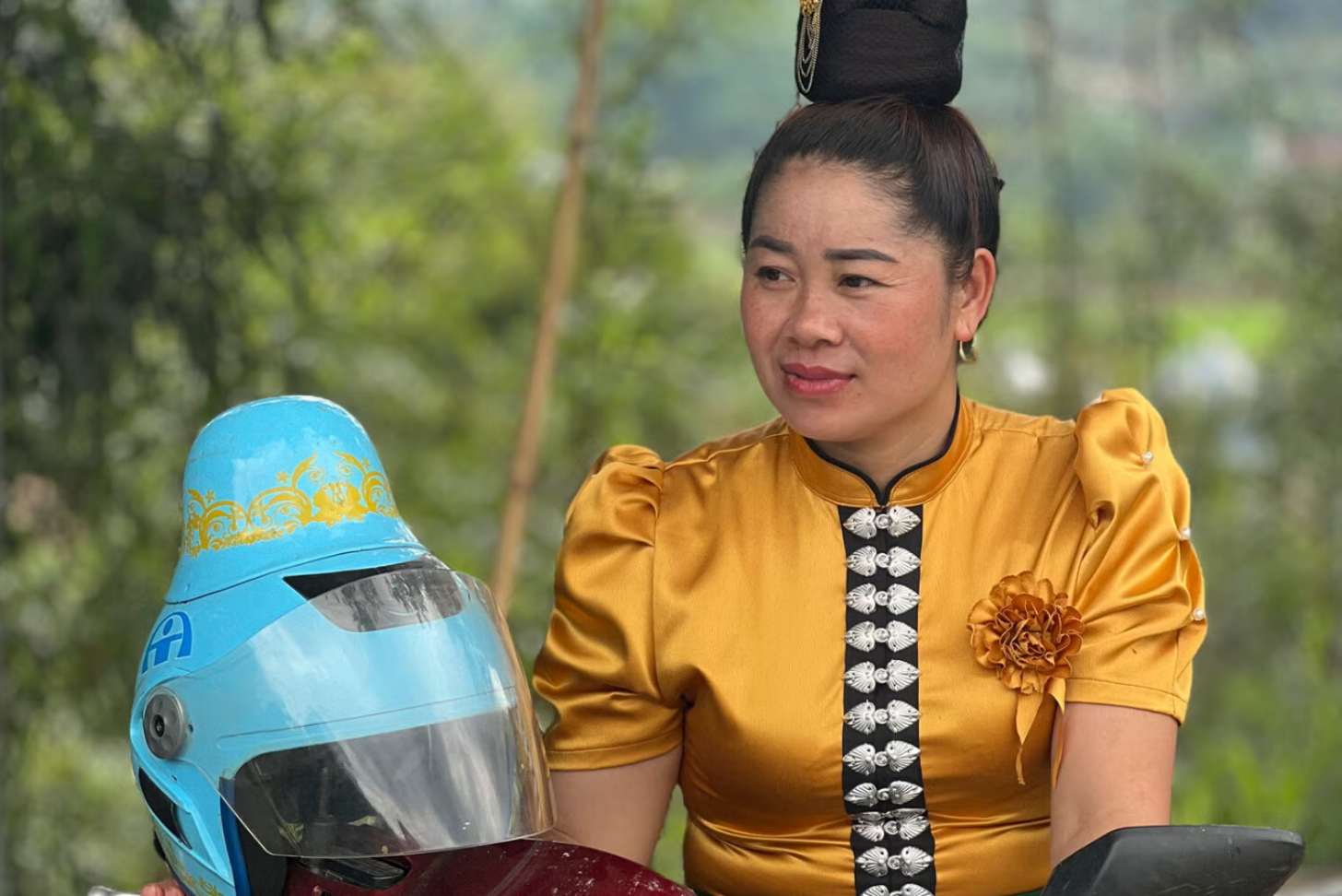 A Thai woman with a high bun sits astride a motorcycle. Beside her is a blue motorcycle helmet with a bump to accomodate buns.