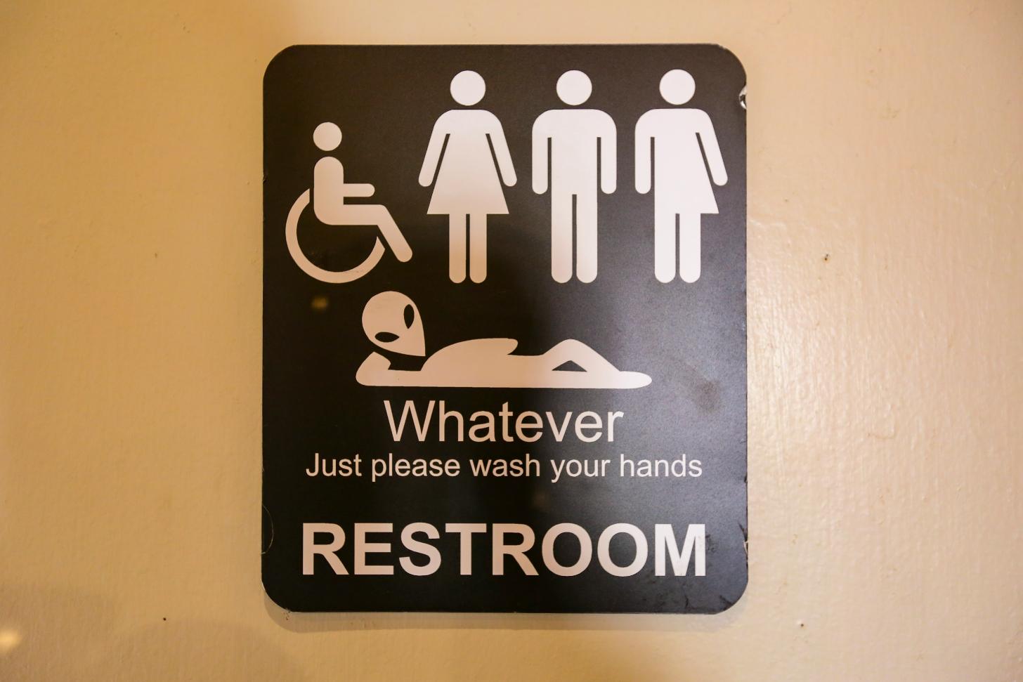 A black sign with white symbols of humans reads "Whatever. Just please wash your hands. Restroom."