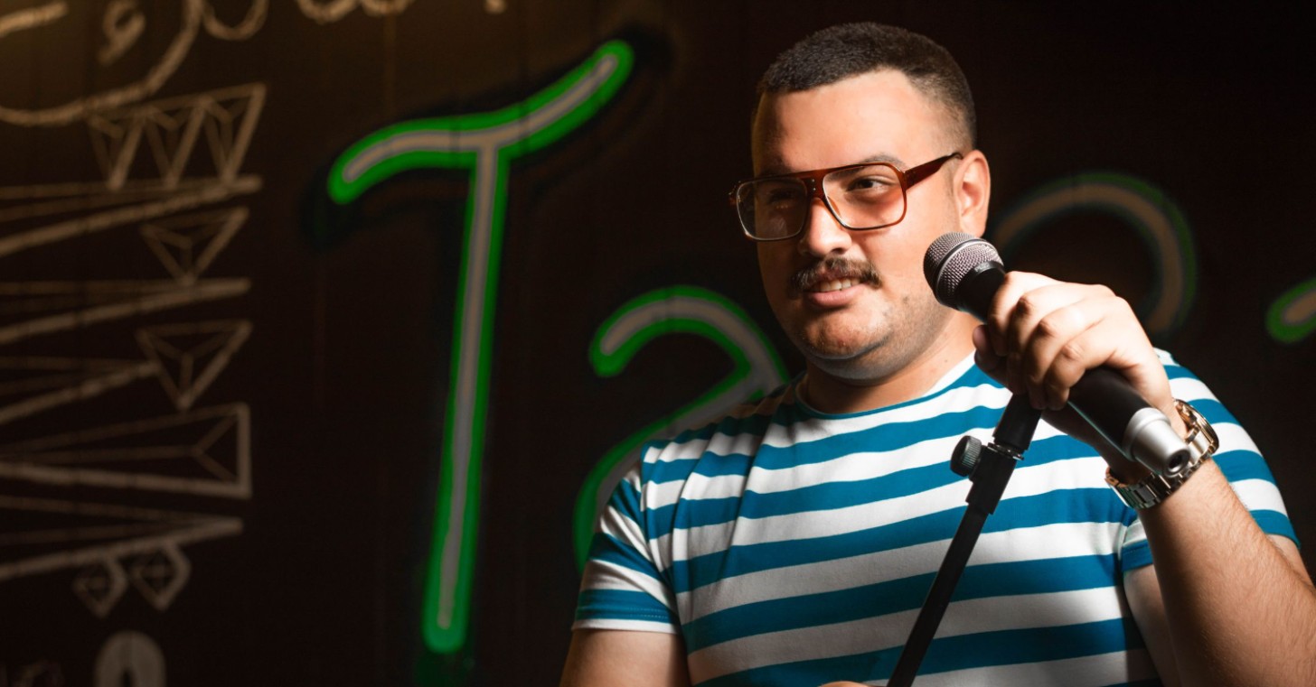 A man with a mustache smiles on a stage as he holds a microphone in his left hand