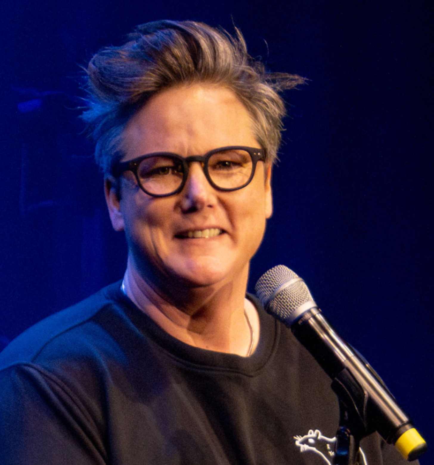 Hannah Gadby, middle aged comedian, smiles from a comedy club stage