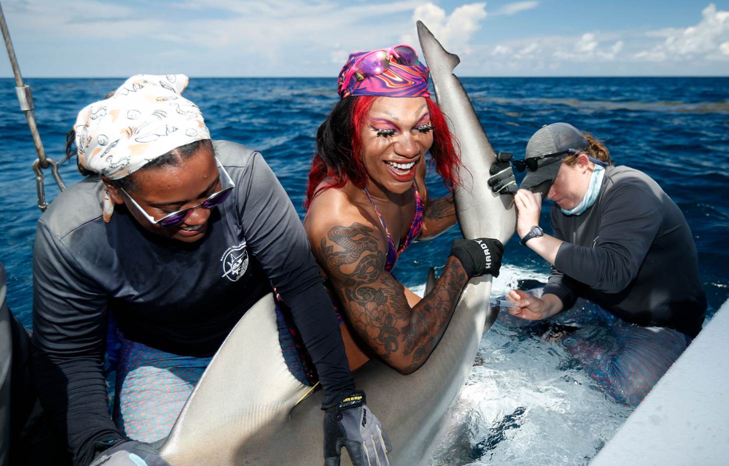 Drag queen Miss Toto smiles as she holds up a massive shark on a boat with the help of two other people.