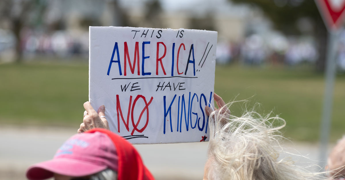 A person holds up a protest sign reading: "This is America; we have no kings!"