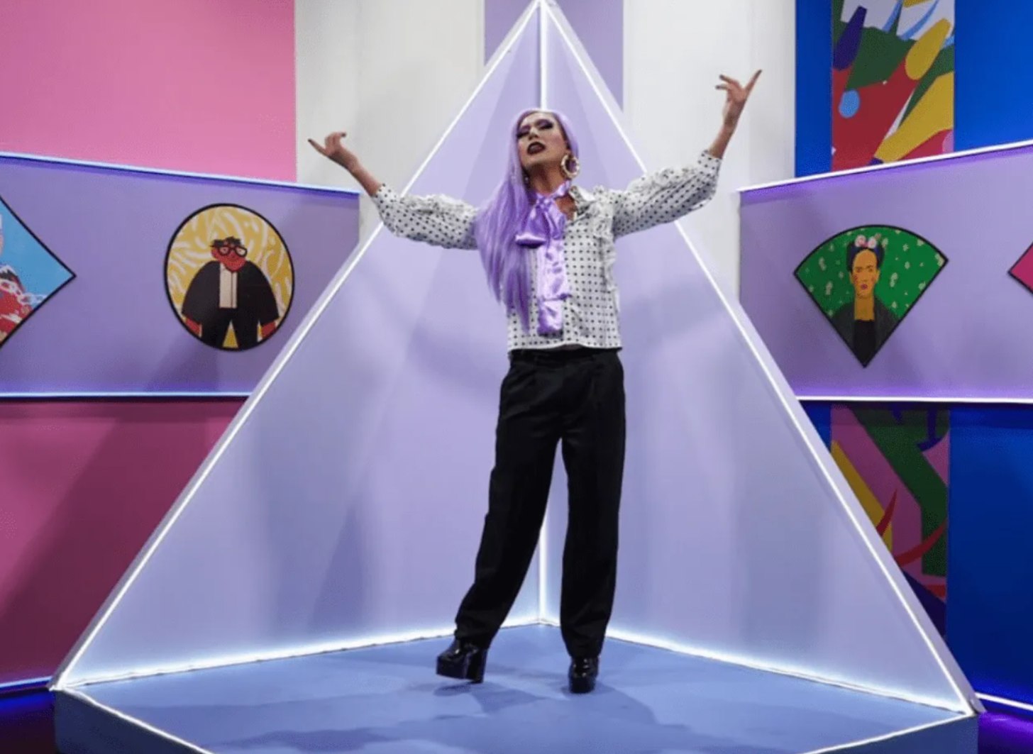 Amanda Drag, a drag queen newscaster, stands on a television stage with her arms outspread.
