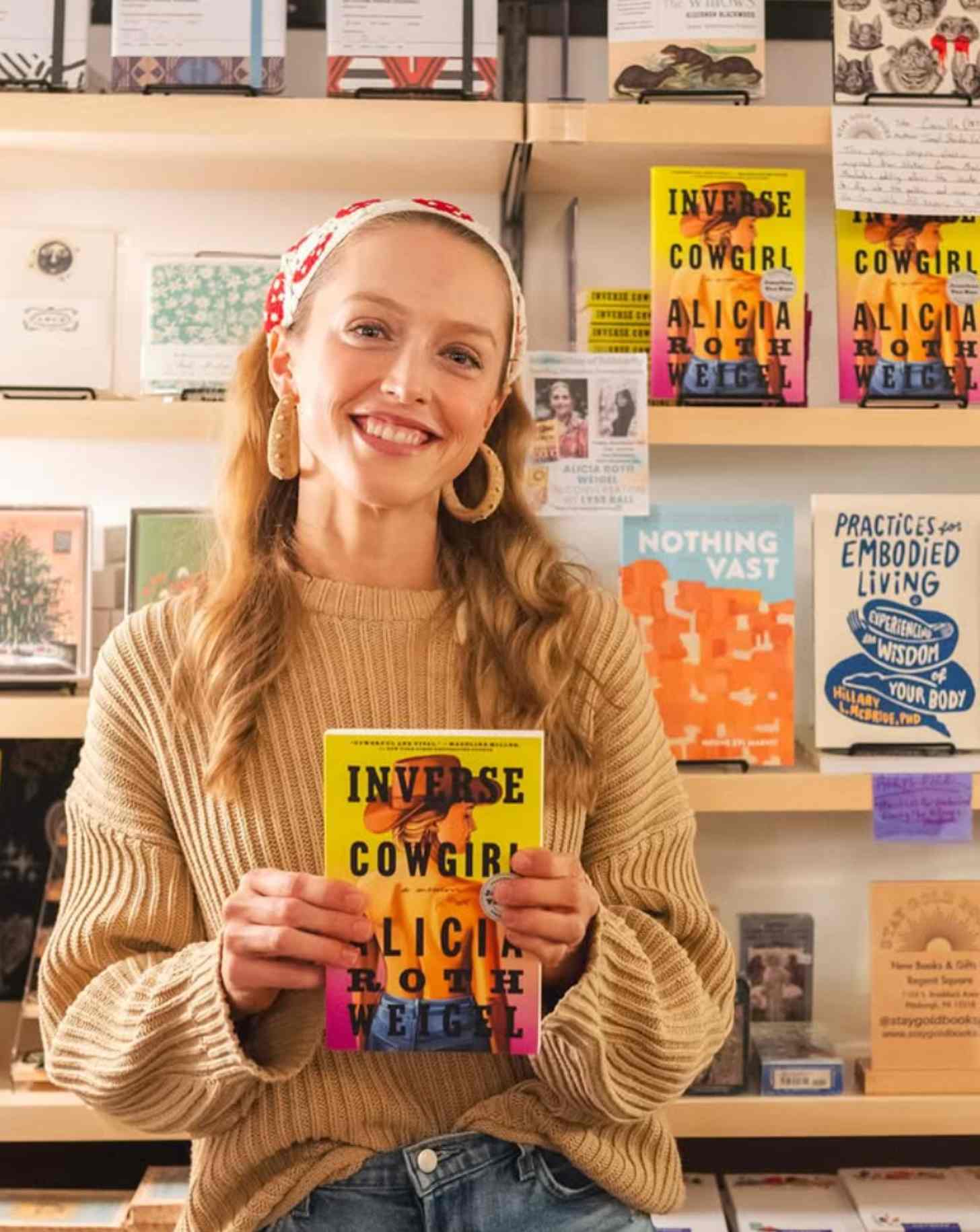 A young blonde woman, Alicia Roth Weigel, stands in a bookstore holding her book: "Inverse Cowgirl"