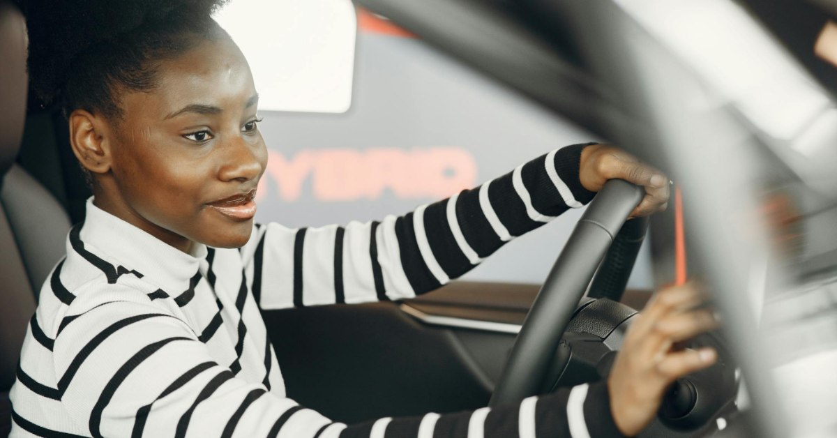 A young Black female driver in a striped shirt smiles as she drives a car