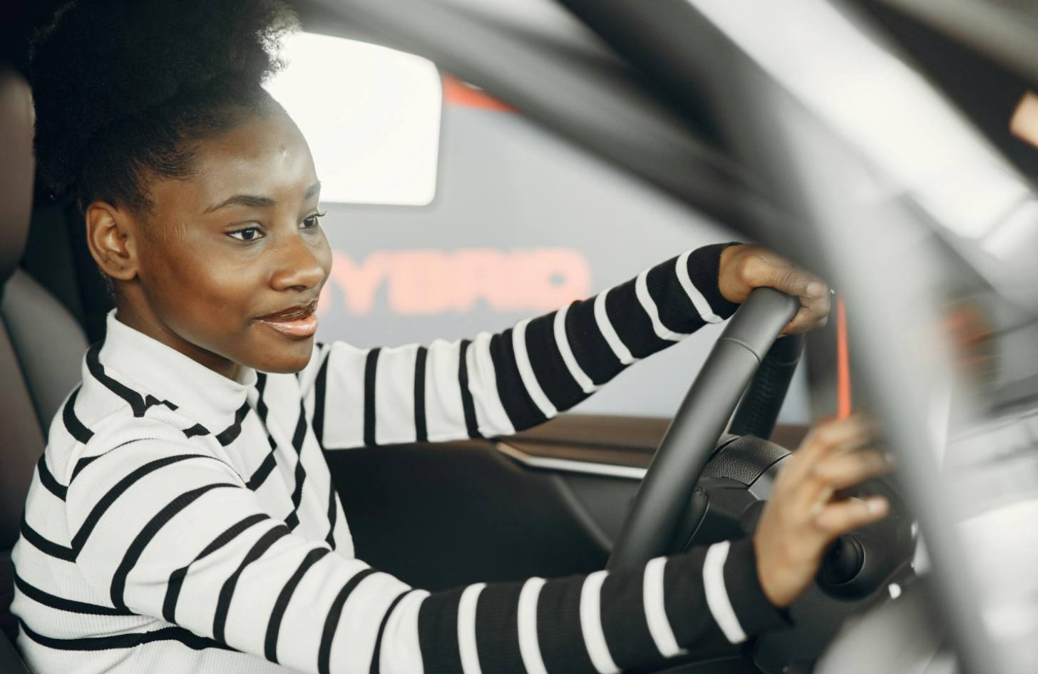 A young Black female driver in a striped shirt smiles as she drives a car