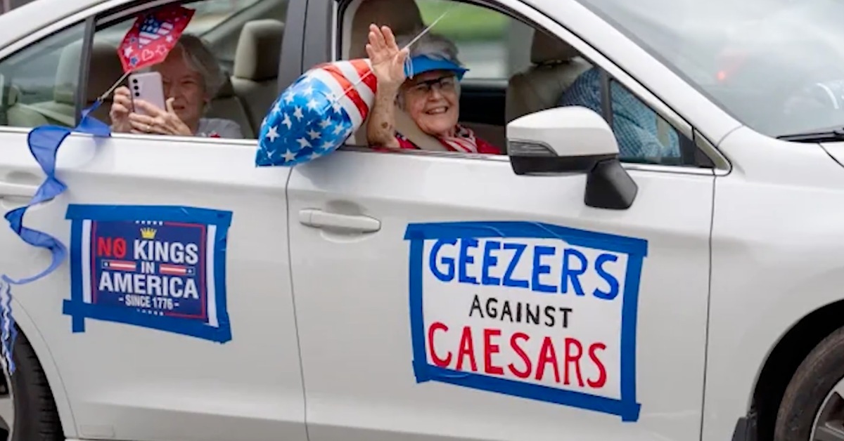 Smiling elderly ladies wave from a car window. A sign taped to their car says "Geezer Against Caesars"