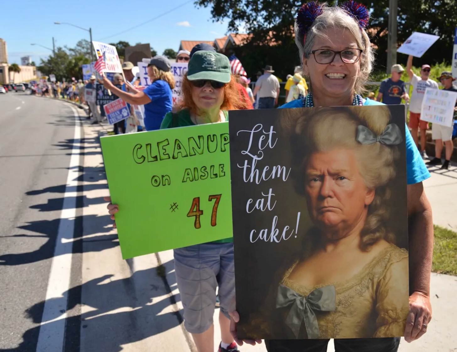 A smiling woman holds up a sign that says: Let Them Eat Cake, with Trump redesigned to look like Marie Antoinette. Beside her, another woman holds a sign that says "Cleanup on Aisle 47"