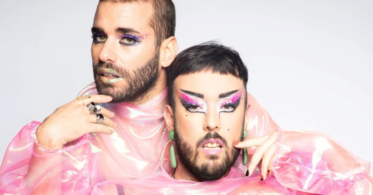 Two bearded musicians with fabulous makeup and pink dresses pose for a photoshoot against a blank canvas