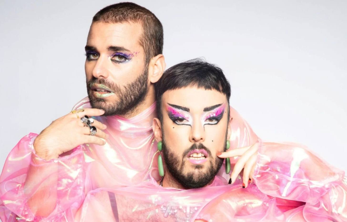 Two bearded musicians with fabulous makeup pose for a photoshoot against a blank canvas