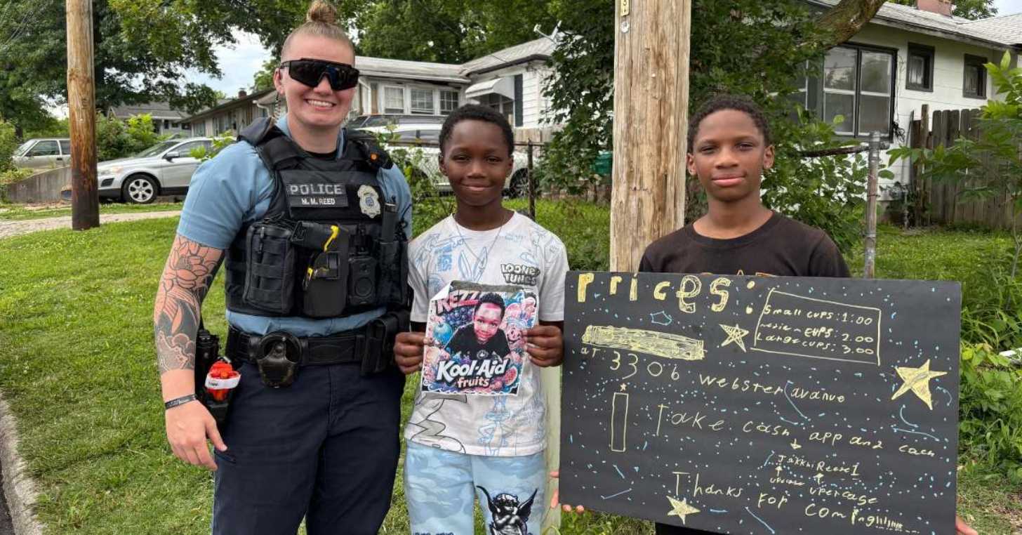 A female police officer smiles as she stands next to two young black children, one of which is holding up a menu for a lemonade stand